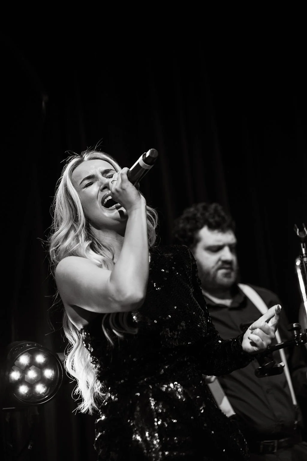A black and white photo of a woman singing passionately into a microphone on stage, with a man playing the violin in the background.
