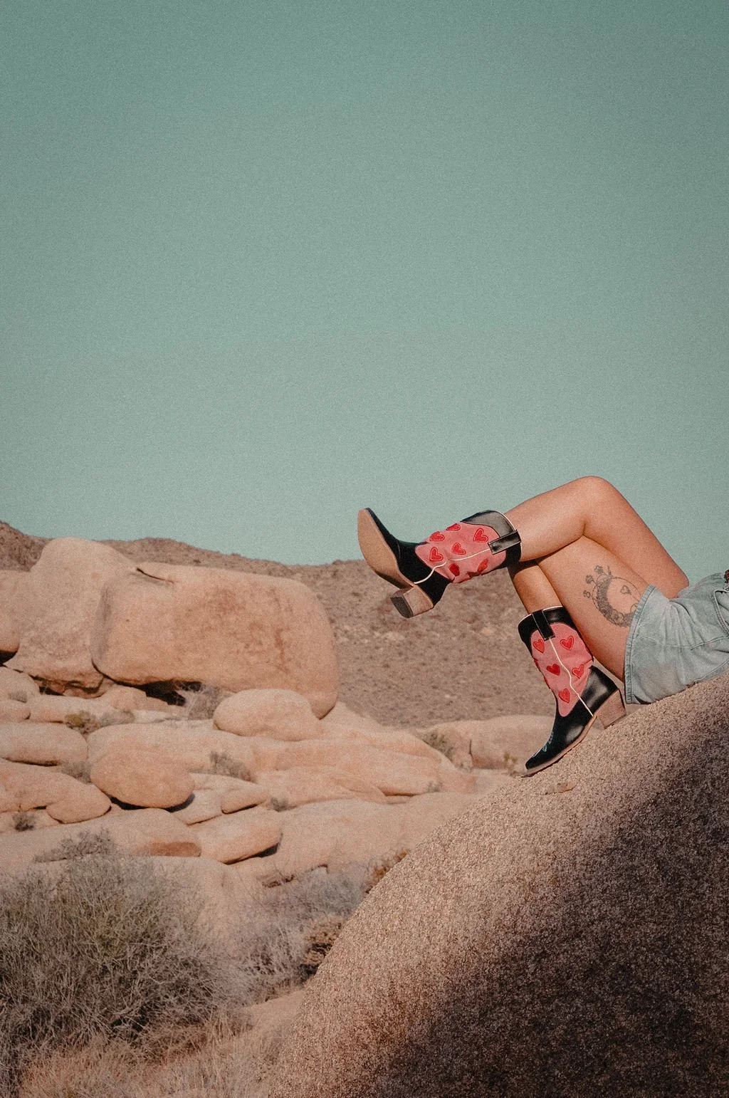 A person is lying on a large rock in a desert landscape with rocky hills in the background. They are wearing black high-heeled boots with pink heart and sock details, denim shorts, and have a tattoo on their thigh.