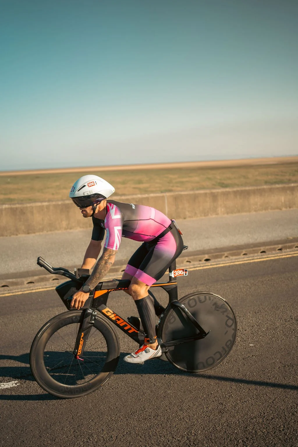 Man riding a black and orange racing bicycle with a disc wheel in a pink and black cycling suit, white helmet, and sunglasses on a road with flat landscape and a clear sky.