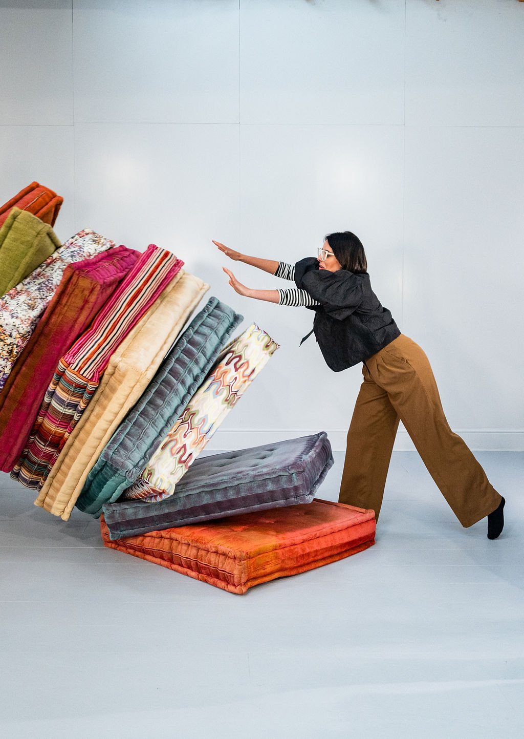 A woman in brown pants, a black blazer, and striped shirt pushes a large stack of colorful cushions and pillows over.