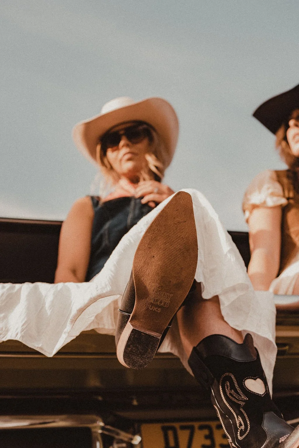 A woman wearing a large sunhat, sunglasses, and a dress sitting at a table with her foot extended toward the camera, showcasing her shoe and the underside of her foot.