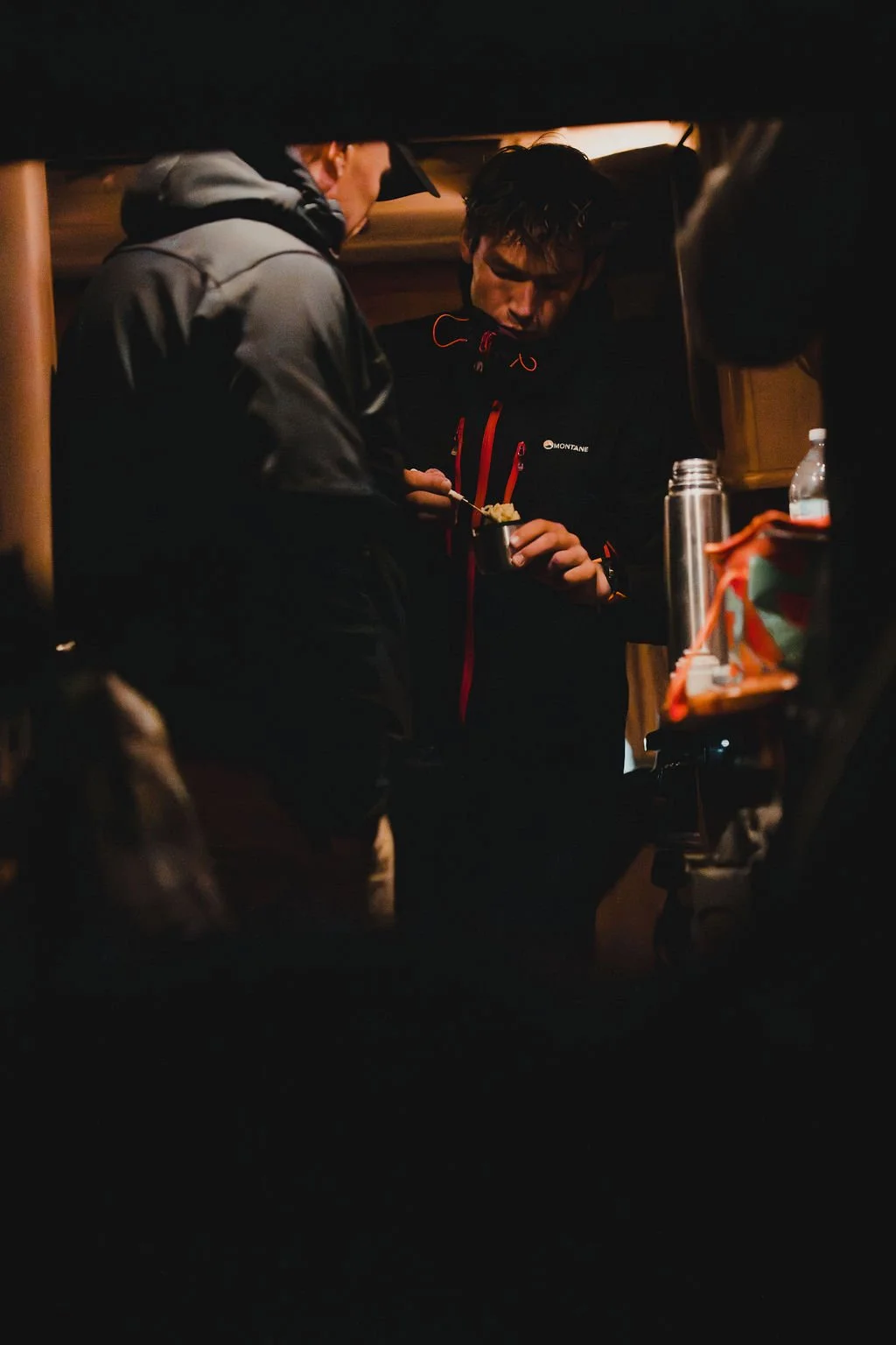 Two men in outdoor jackets inside a vehicle, one holding a bowl and spoon, the other looking at it, with bottles and a thermos on a small table.