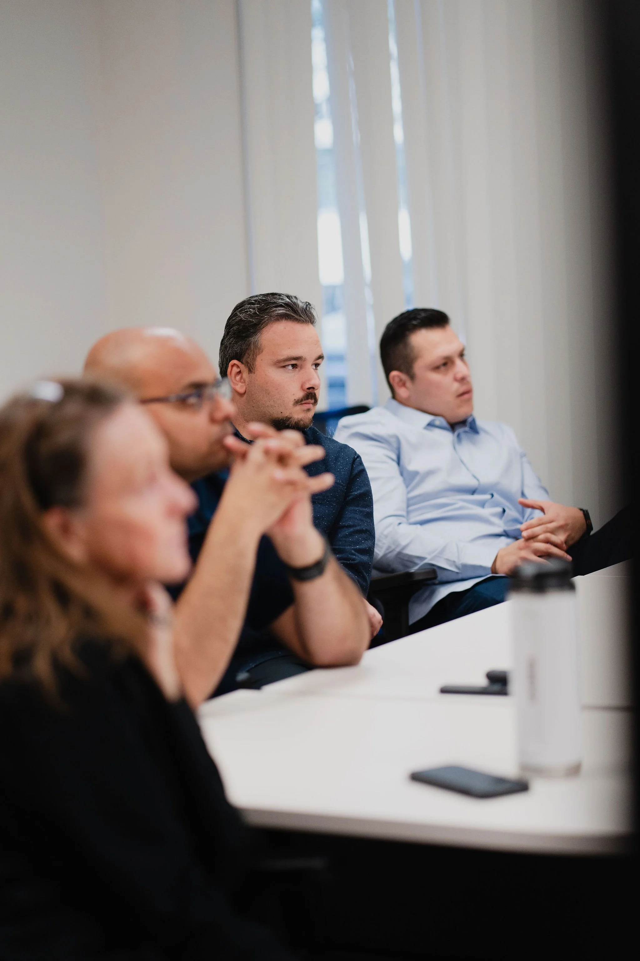 A group of four professionals attending a meeting or seminar, sitting at a conference table, focused on a presentation or speaker, with a white wall and window behind them.