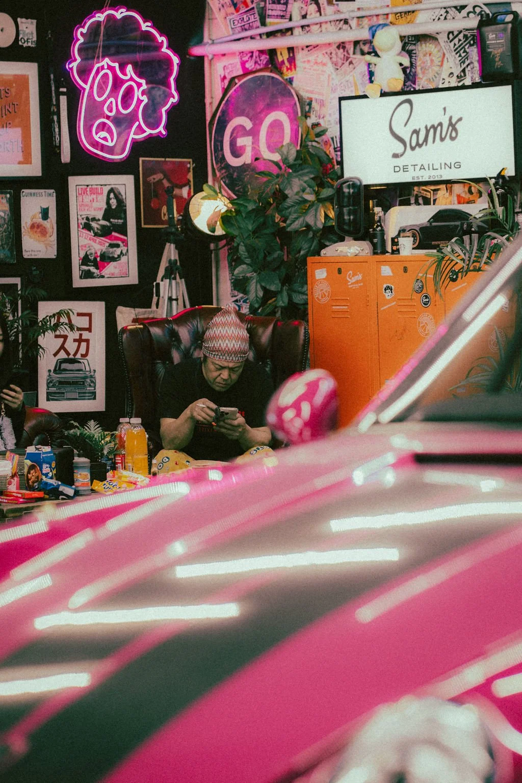 Akira Nakai wearing a patterned headscarf and black shirt sitting on a large black leather chair, looking at his phone inside a colourful, eclectic space decorated with neon signs, posters, plants, and lockers.