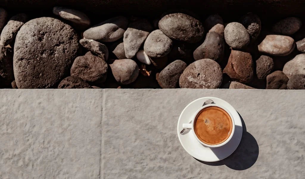 A top view of a cup of coffee on a saucer placed on a stone surface outdoors, with rocks in the background.