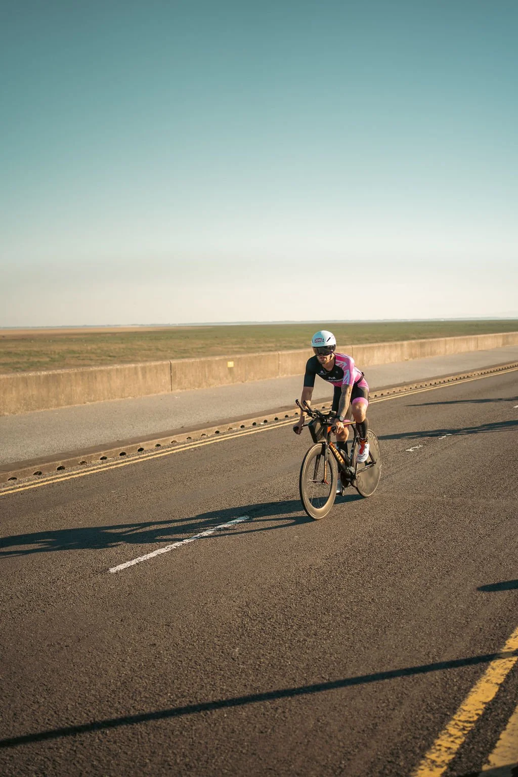 A cyclist wearing a helmet and sunglasses riding a black and orange bike on an open road with a grassy field and clear sky in the background.