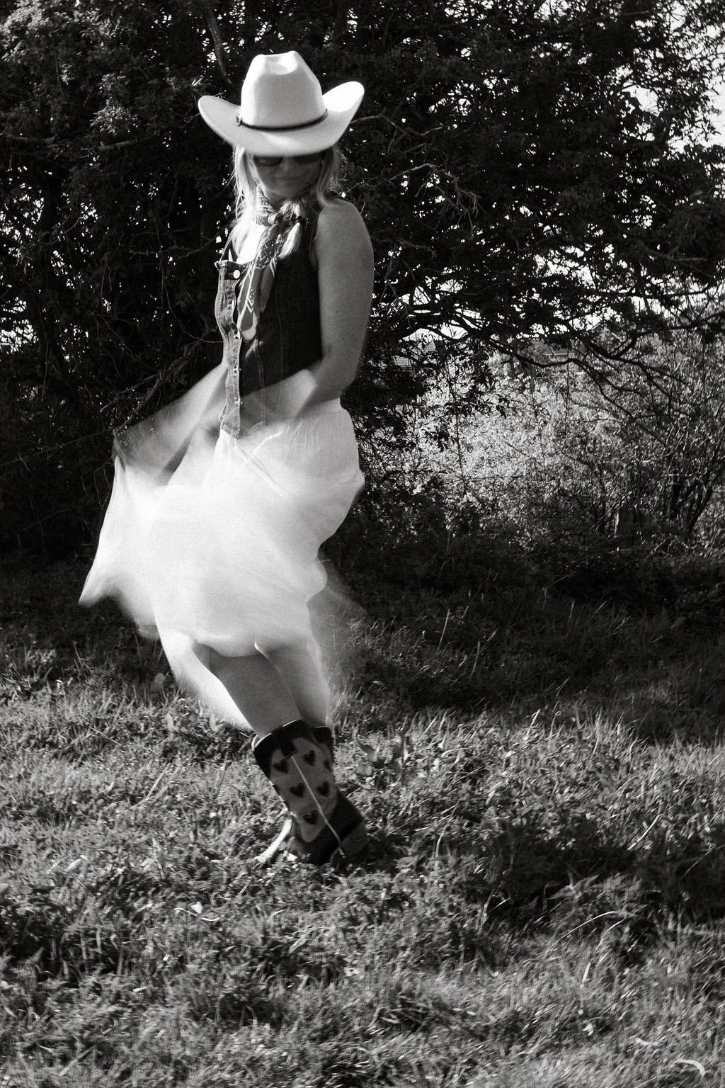 A woman wearing a cowboy hat, sunglasses, a sleeveless top, a skirt, and cowboy boots with heart patterns, is standing outdoors on grass near bushes or trees, with her head slightly bowed and hands touching her skirt.