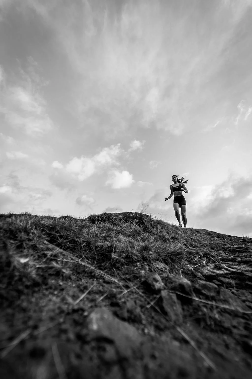 A woman running on a grassy hilltop outdoors with a cloudy sky in background, captured in black and white.
