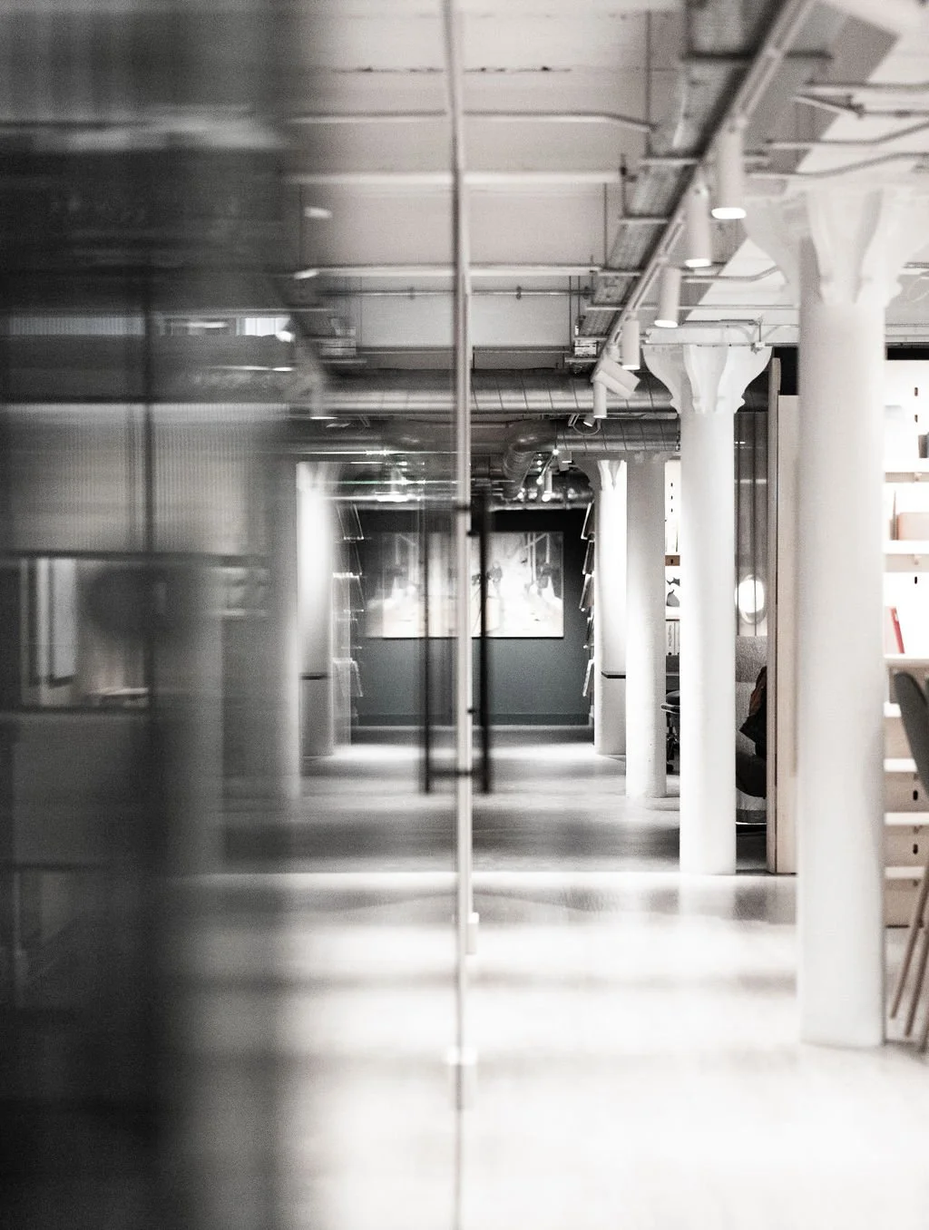 Empty modern interior corridor with white columns, exposed ceiling pipes, and a glass reflection on the left side, leading to a faint distant view.
