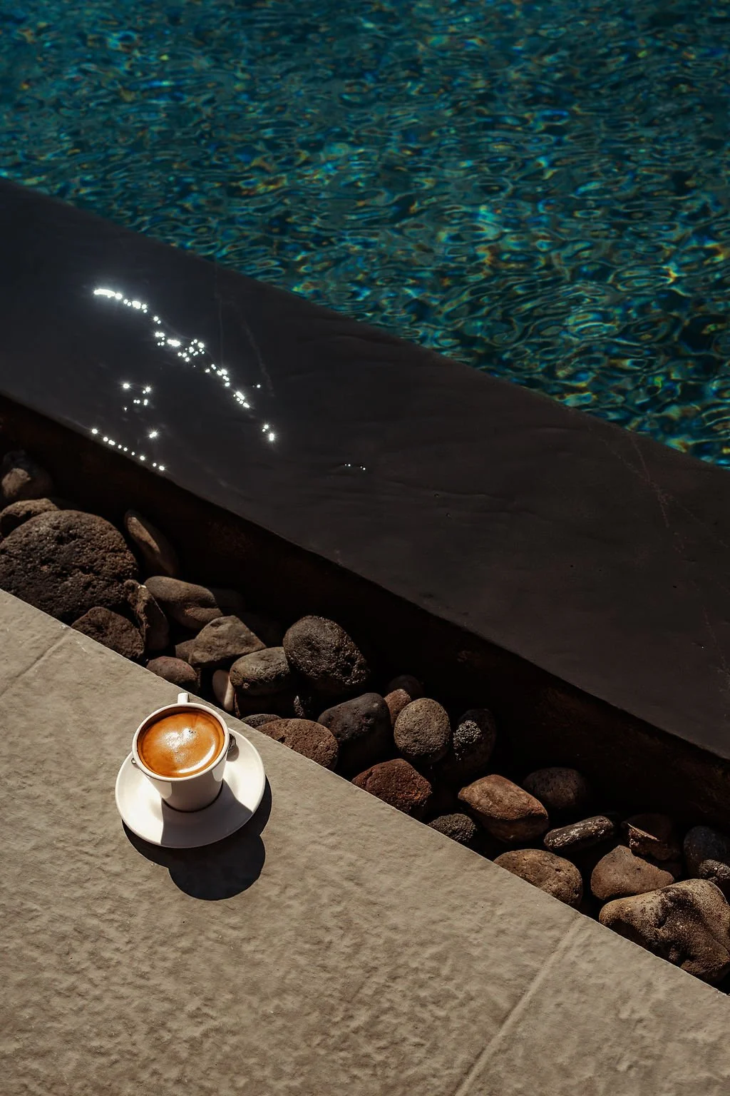 A cup of coffee on a white saucer resting on a light-colored stone surface near a pool with dark black stone lining, surrounded by small rocks. The pool water is blue with reflections of light.