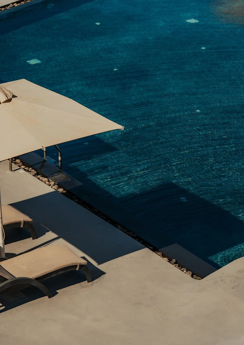 Empty swimming pool with blue water, beige umbrellas, and lounge chairs on a concrete poolside deck.
