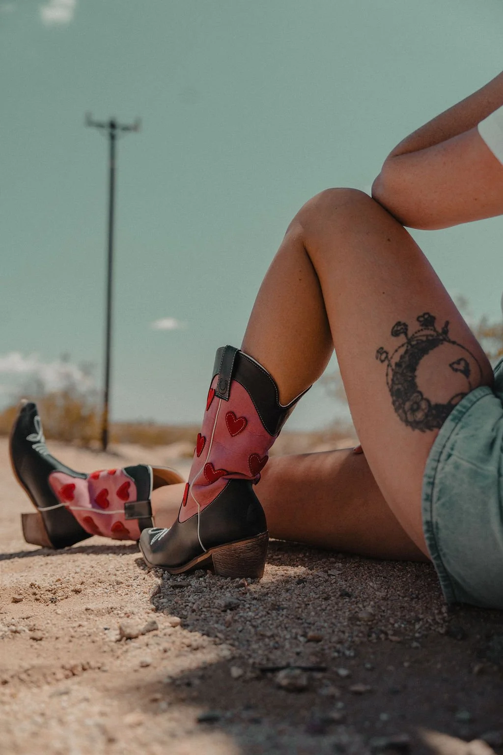 A person sitting on sandy ground wearing cowboy boots with red heart patterns and denim shorts, with a visible tattoo on the thigh, under a clear sky with a utility pole in the background.