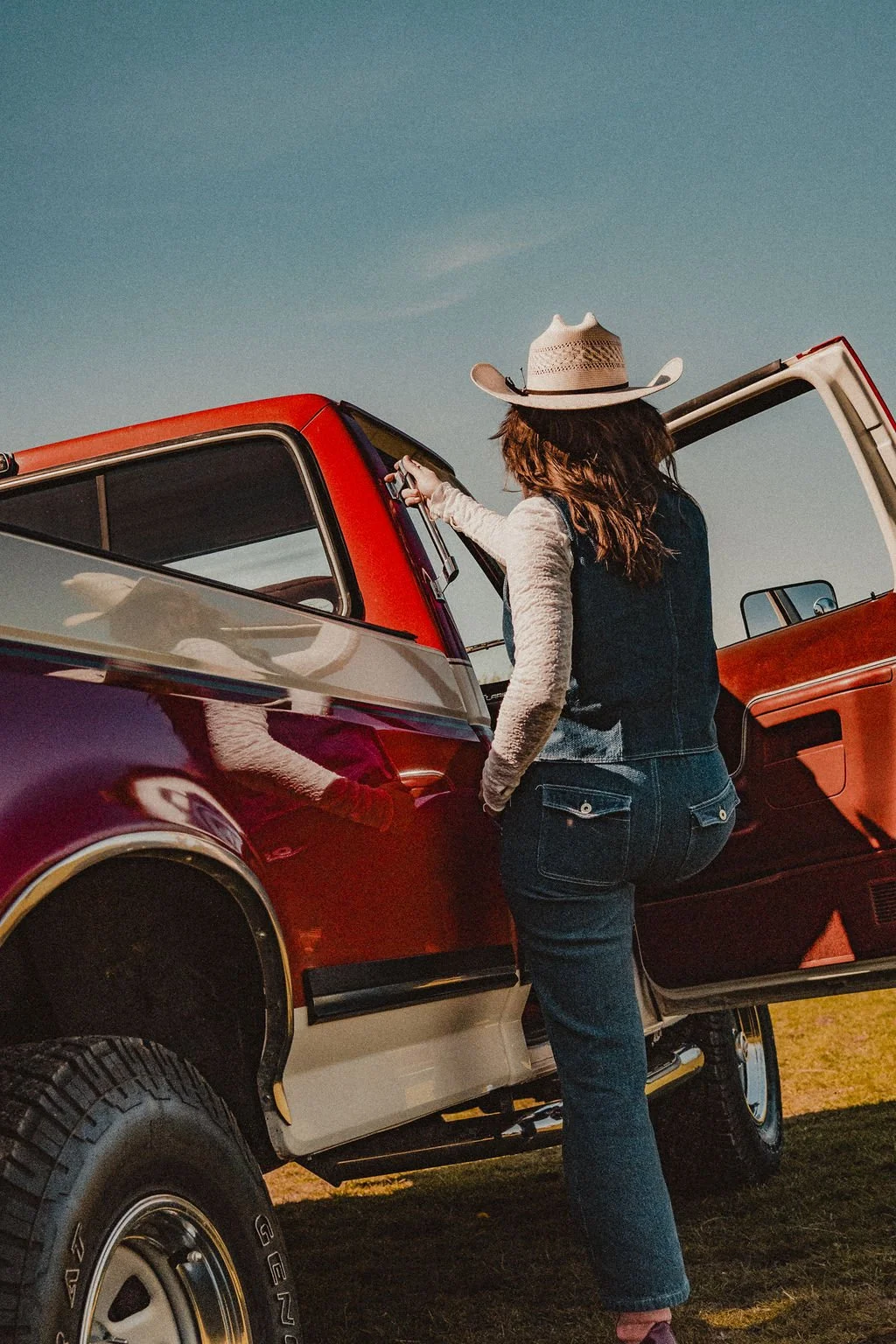 A woman with long hair wearing a cowboy hat, denim vest, and jeans, standing next to a red and white pickup truck with an open door, in a rural outdoor setting.