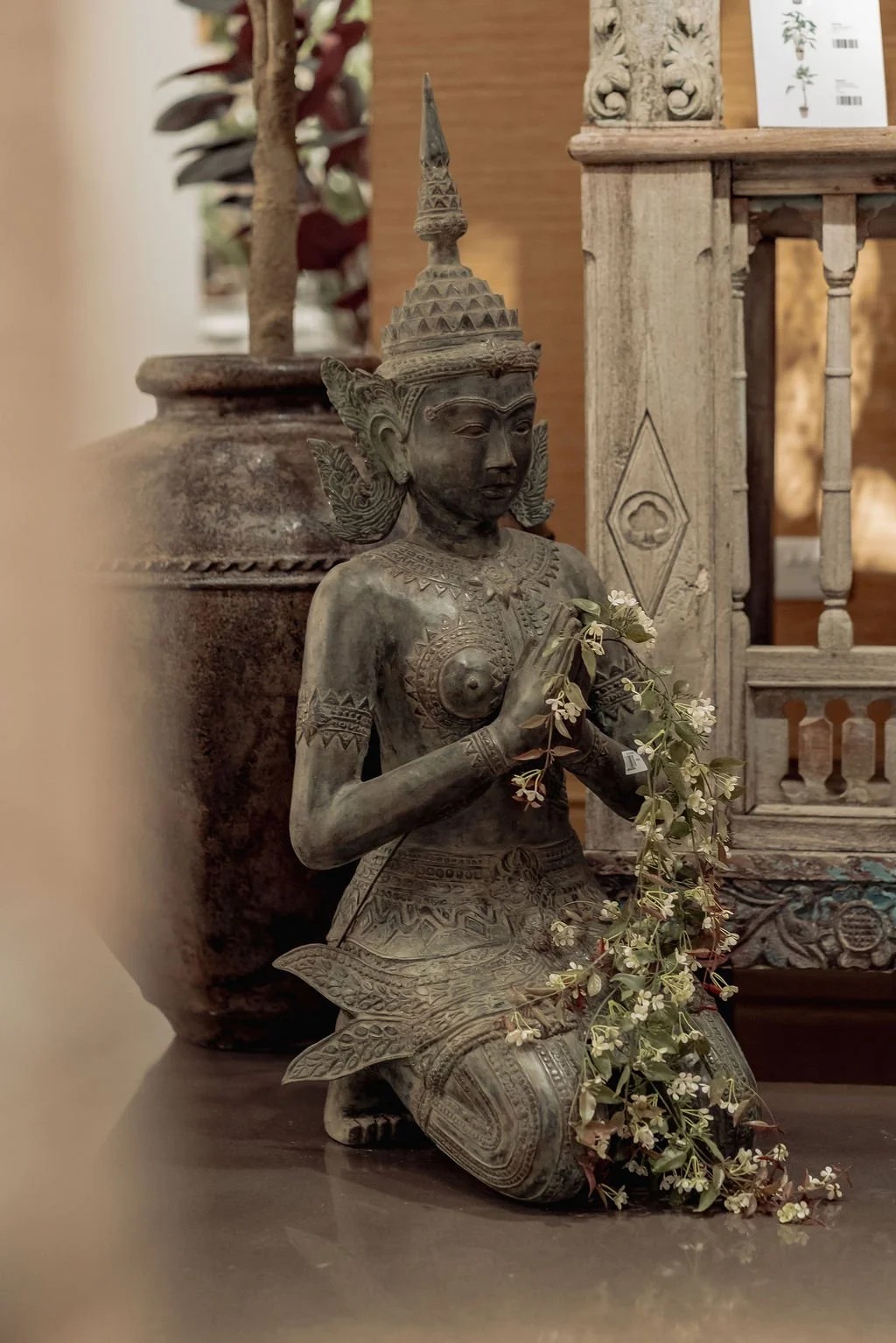 A kneeling statue of a traditional Southeast Asian female deity or figure, made of carved stone, with hands in a prayer or greeting gesture, holding a small branch with white flowers. The figure wears an intricate headdress and jewelry, surrounded by