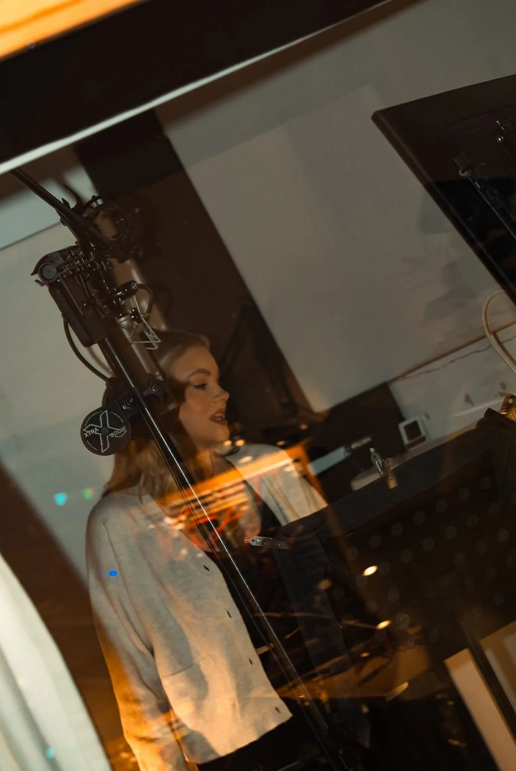 A woman with blonde hair and makeup is singing or speaking into a microphone inside a recording studio, viewed through a glass window.