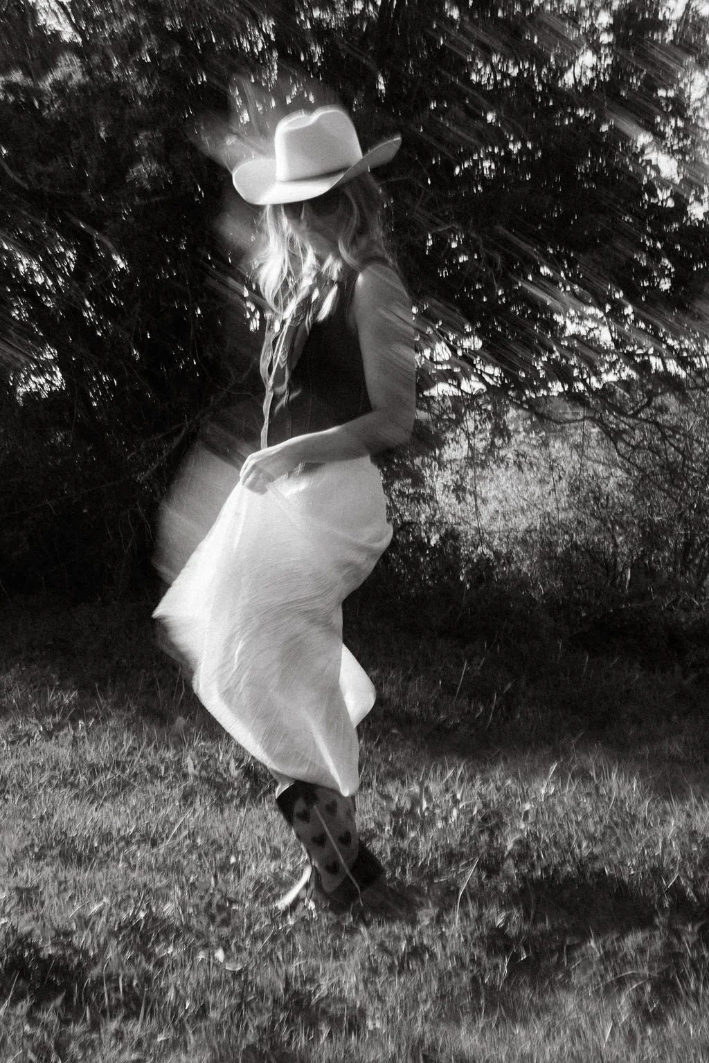 A woman in a cowboy hat, long skirt, and heart patterned cowboy boots standing outdoors at night, with trees and grass in the background.