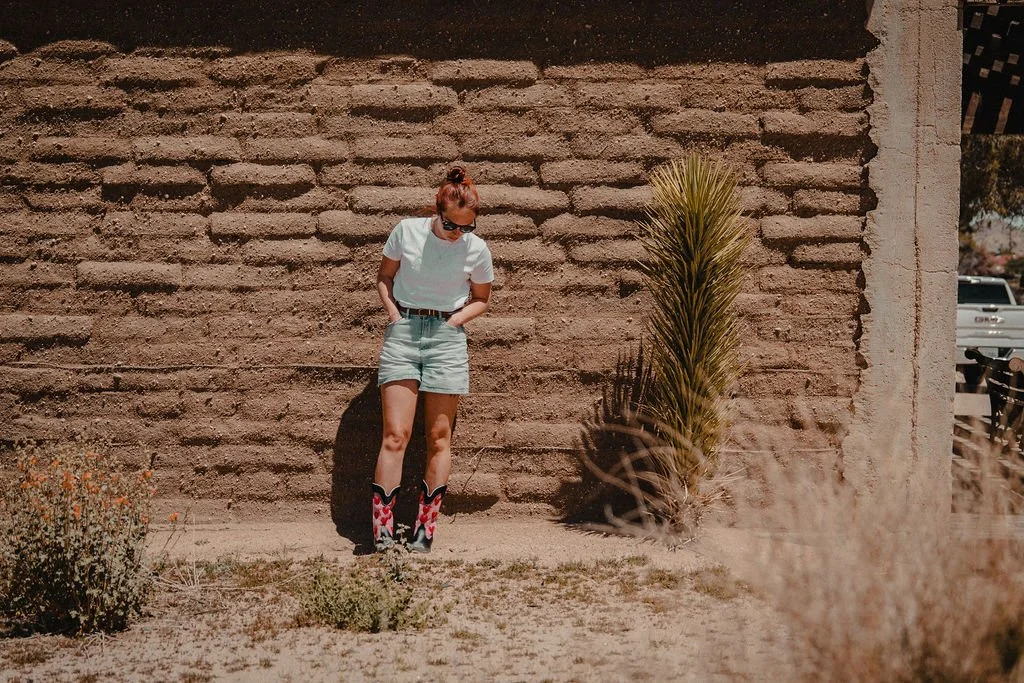 A woman with sunglasses and red hair tied up in a bun, wearing a white t-shirt, high-waisted denim shorts, and cowboy boots, standing outdoors against a rough brick wall and a small palm plant.