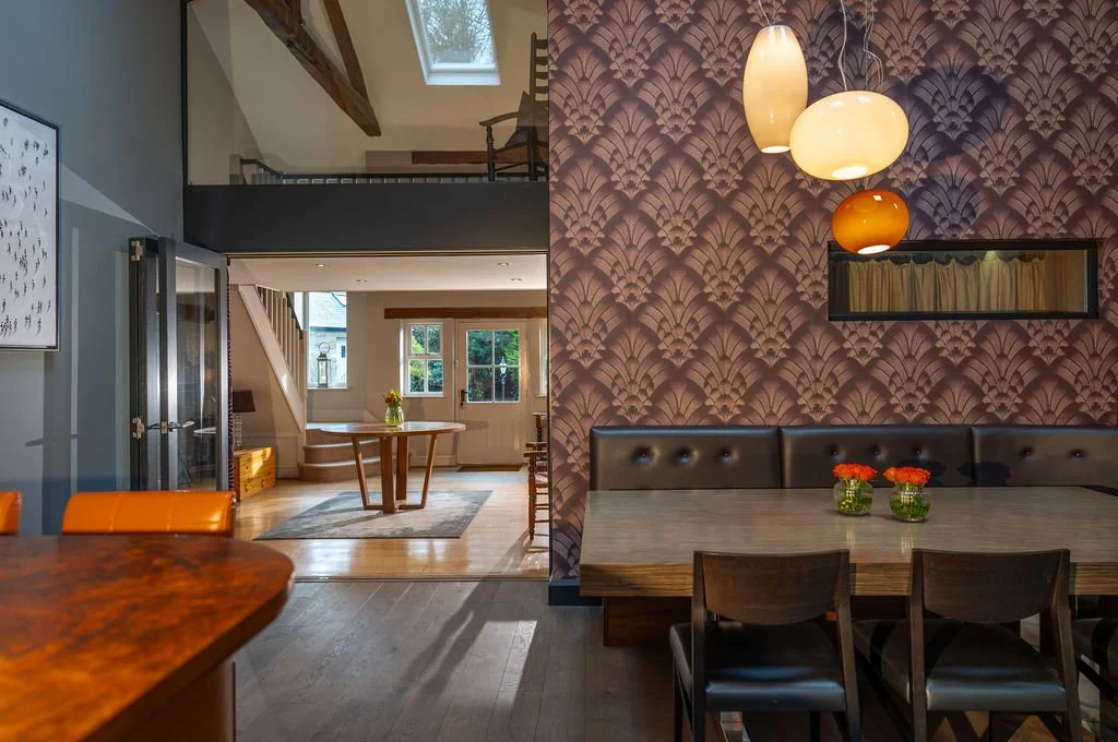 Interior of a home with a dining area and a sitting area, featuring a purple patterned wallpaper, chandelier, and natural light from windows and a skylight.