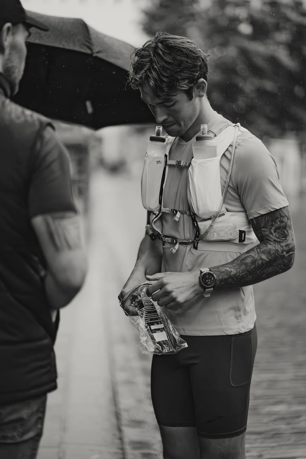 A young man in athletic gear, holding a water packet, with a hydration pack on his chest, standing outdoors during a rainy day, being helped by another person holding an umbrella.