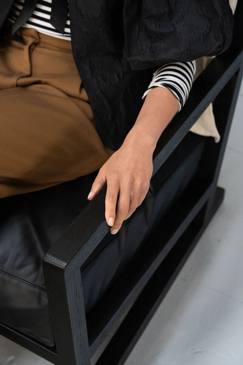 Person sitting on black furniture with hand resting on armrest, wearing a black vest over a black and white striped shirt, and tan pants.