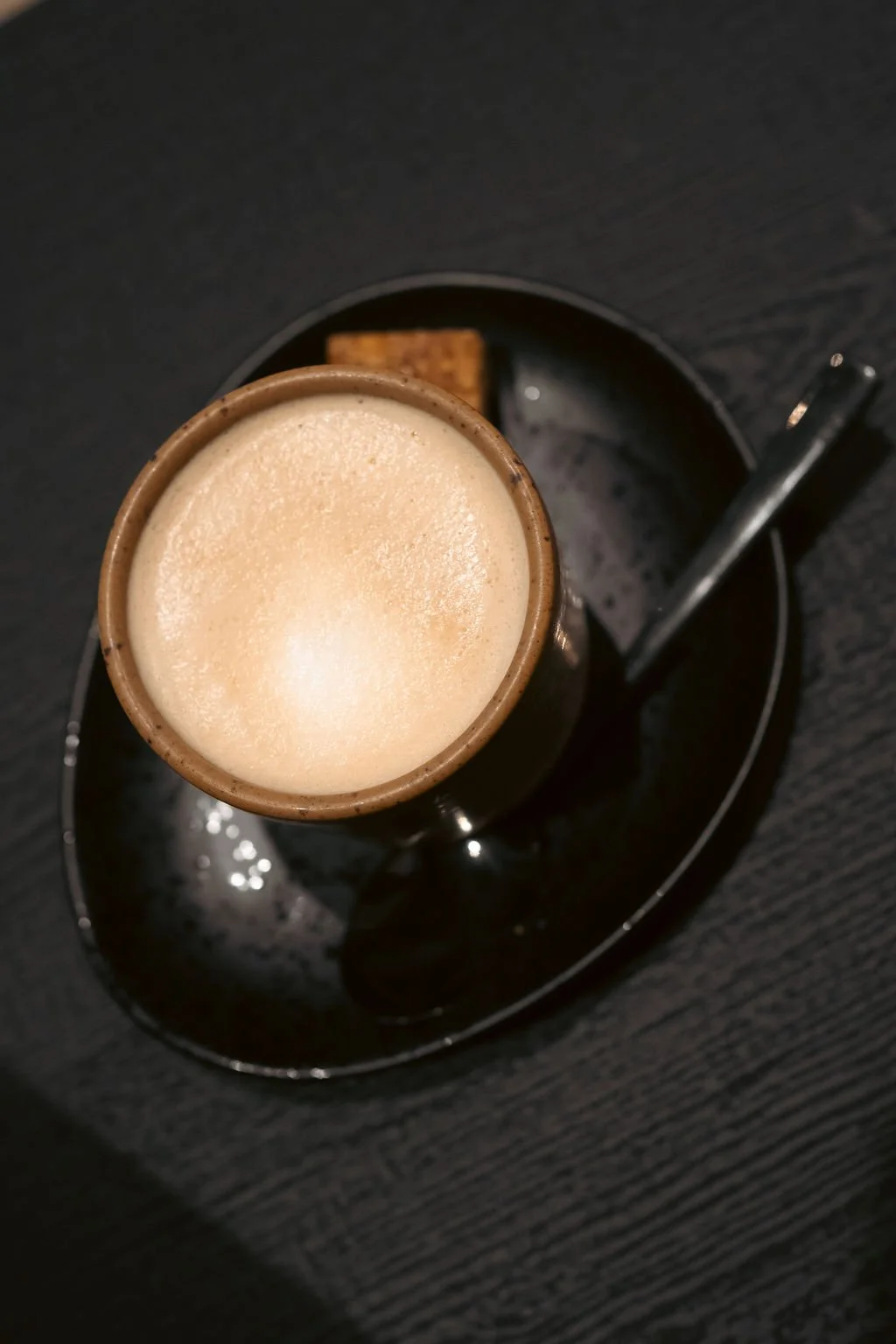 A cup of cappuccino with foam, placed on a small black tray with a chocolate square and a teaspoon, on a dark wooden table.