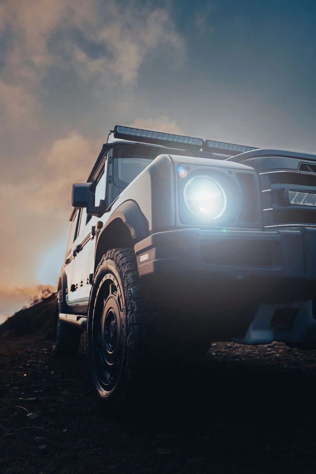 Close-up of a rugged off-road vehicle with its headlight on, driving on a dirt path at sunset or sunrise.