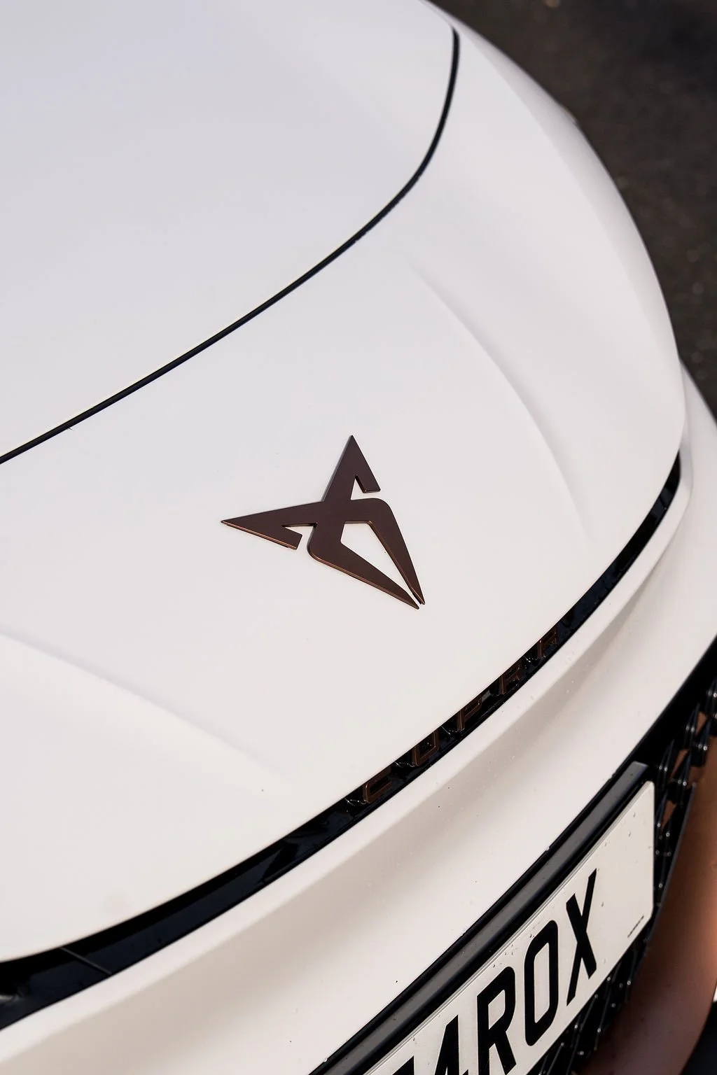 Close-up of a white car's front with a stylized logo and partial license plate reading '4ROX'.