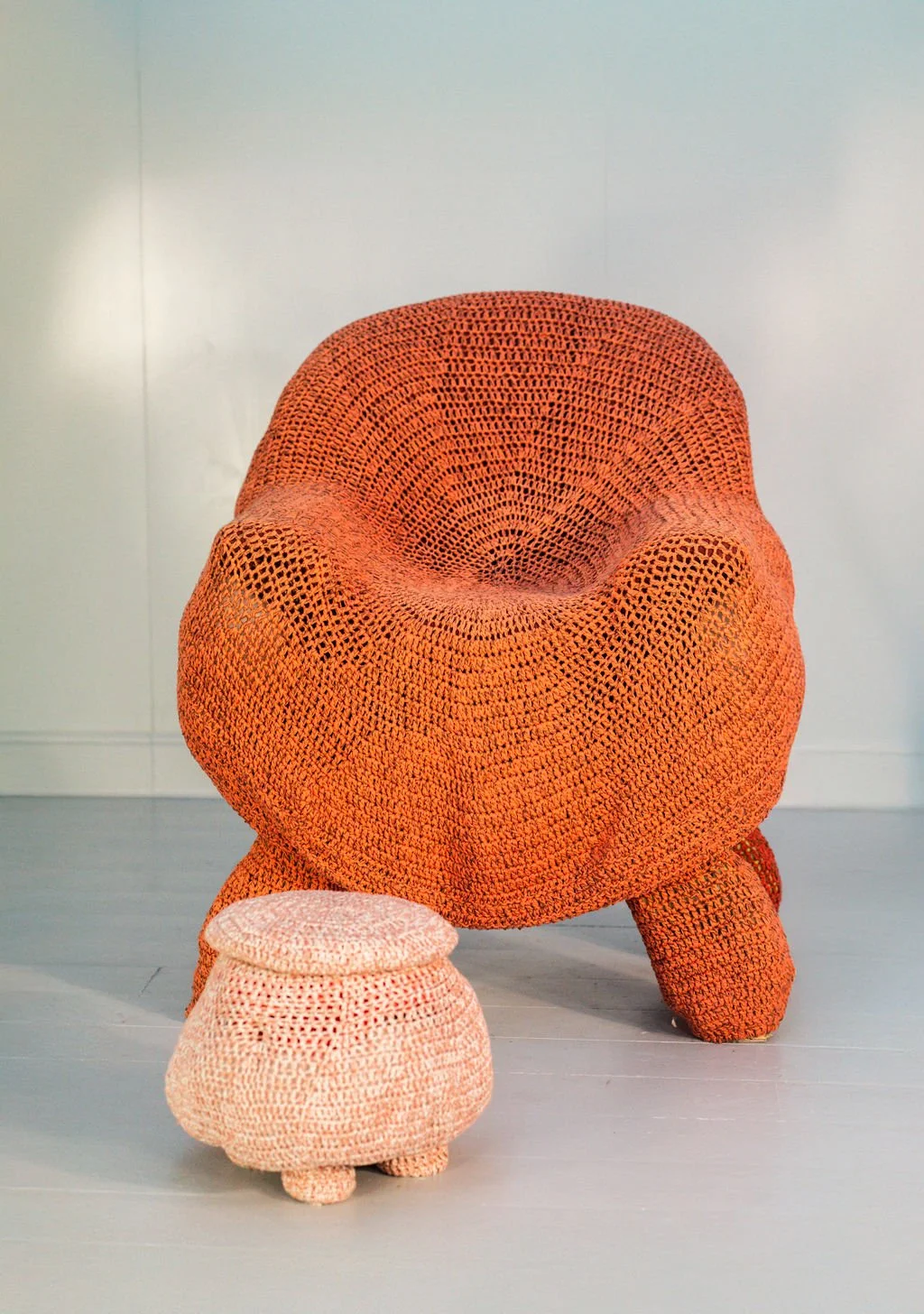 A large, rounded orange chair with a textured, crochet-like surface, positioned next to a smaller matching ottoman in the same style, in a minimalistic room with a light gray floor and plain white wall.
