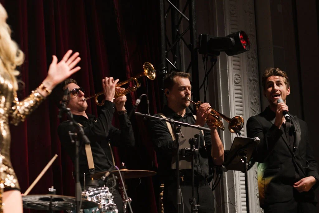 Four musicians performing on stage, with three men playing trumpets and one woman singing into a microphone. The background features dark red curtains and stage equipment.