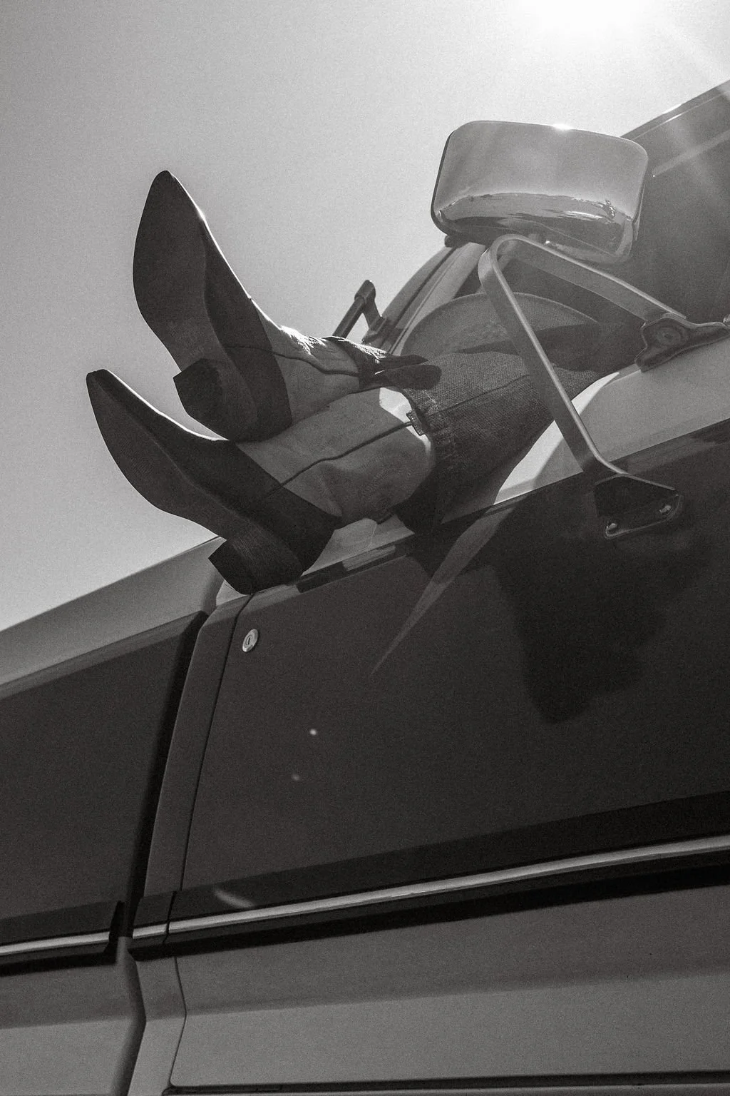 Black and white photo of a person sitting on the hood of a car, wearing cowboy boots, jeans, and a jacket, with the person's legs up and a large side mirror of the car visible.