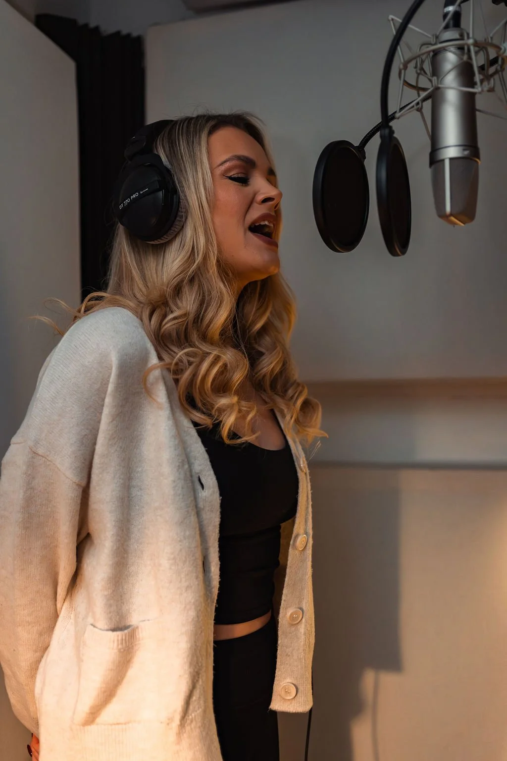 A woman with long curly blonde hair wearing headphones and a beige cardigan, singing into a professional microphone with a pop filter in a recording studio.