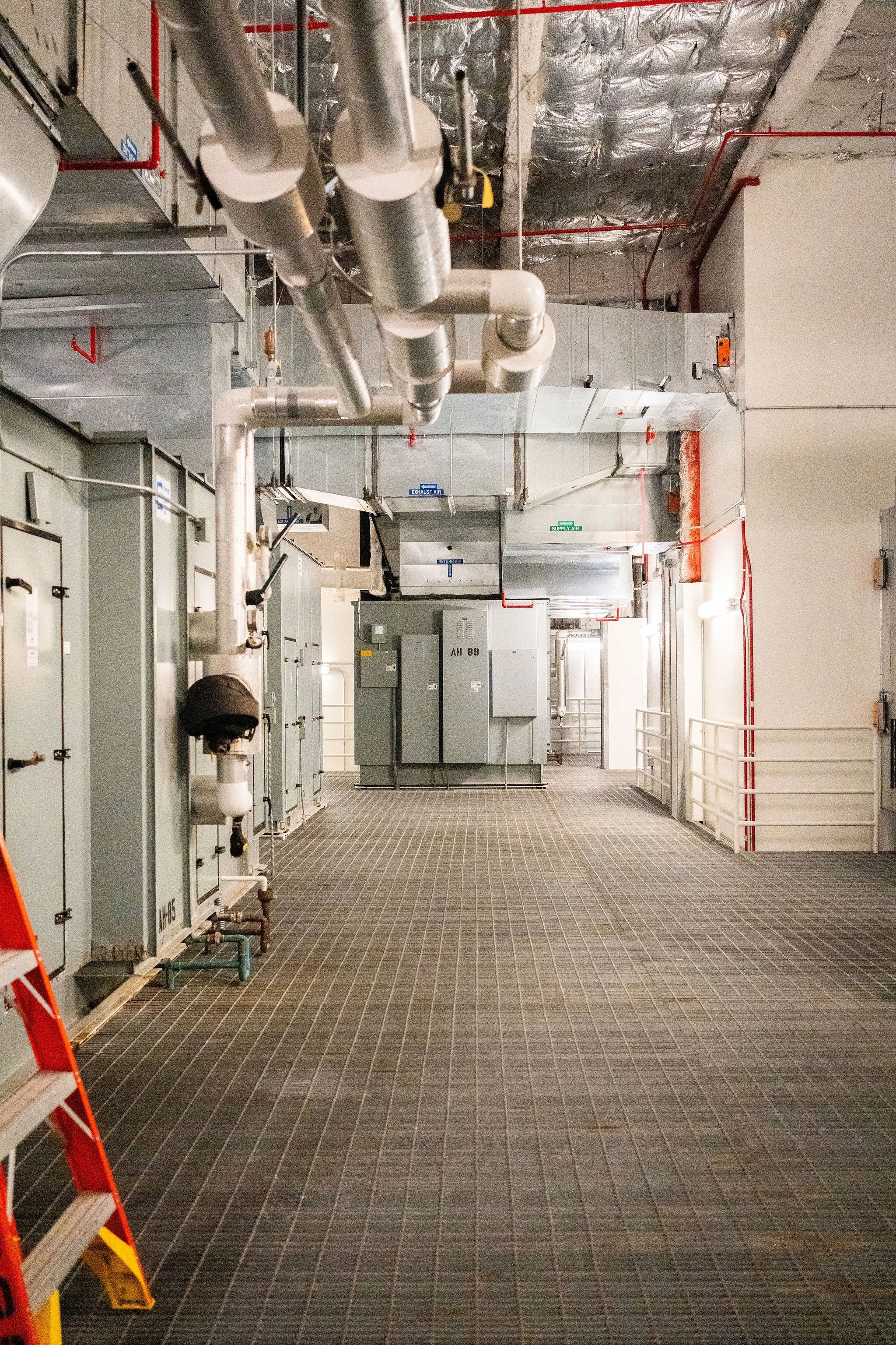 Industrial mechanical room with gray electrical panels, large ventilation pipes, and a metal grated floor.