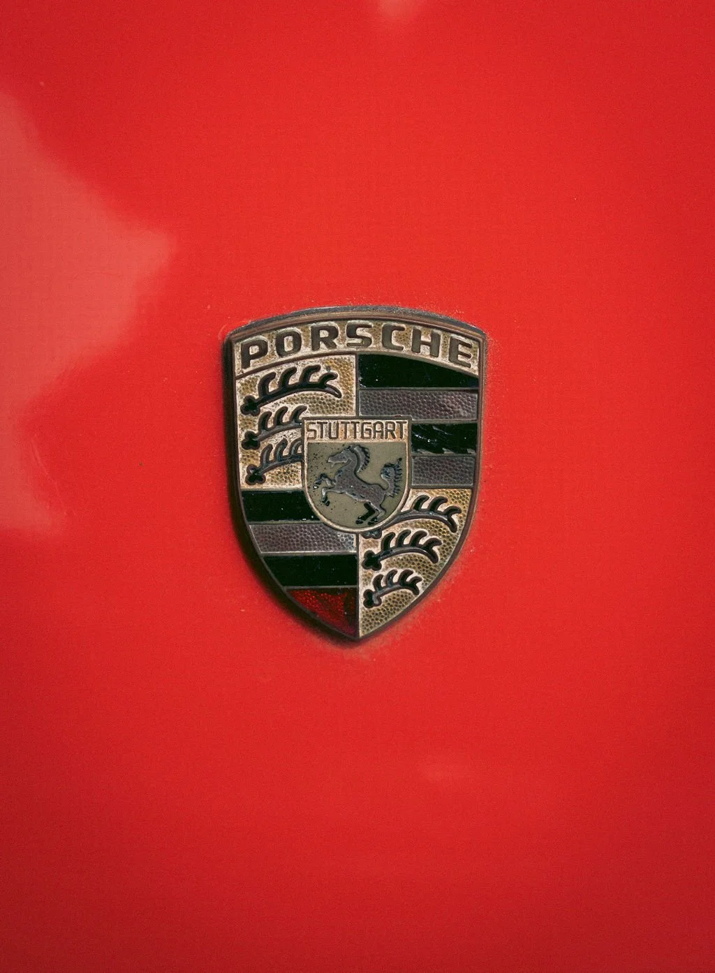 Close-up of a Porsche emblem on a red car, with the word 'Porsche' at the top and a shield with a horse and the word 'Stuttgart' in the center.