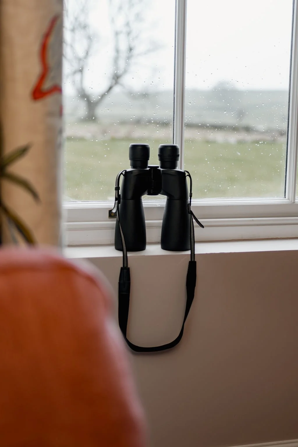 A pair of black binoculars on a windowsill with raindrops on the window and a blurry outdoor landscape in the background.
