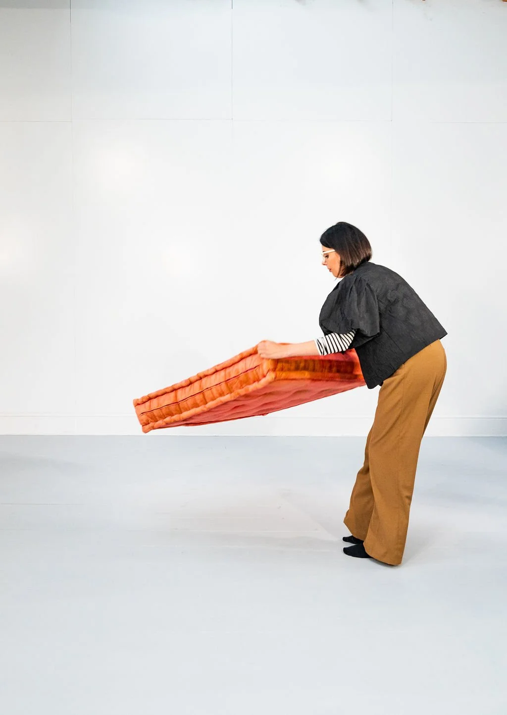 A woman holding a large, orange cushion in a minimalistic white room.