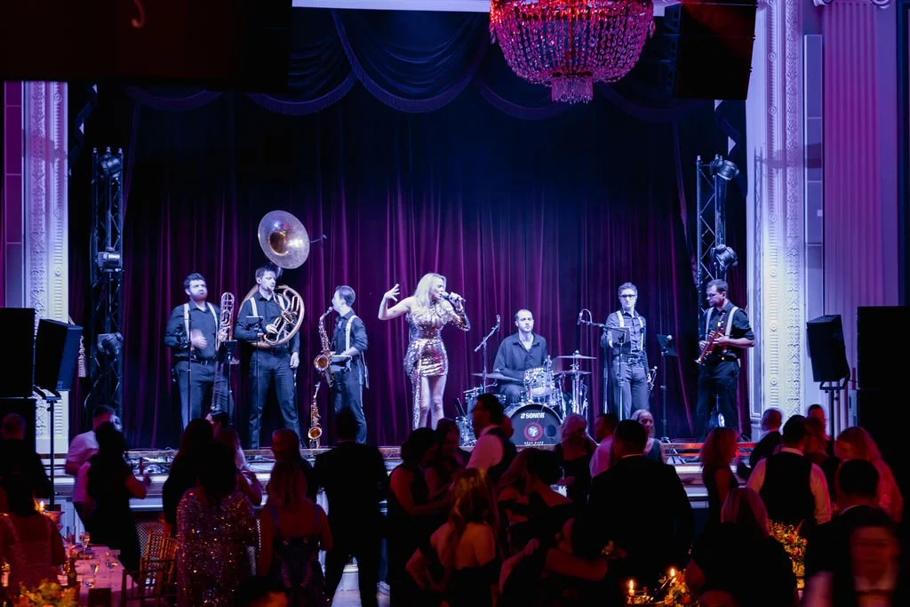 Band performing live on stage at a formal event, people dancing and socializing in the audience, elegant decor with chandelier, dark red velvet curtains.