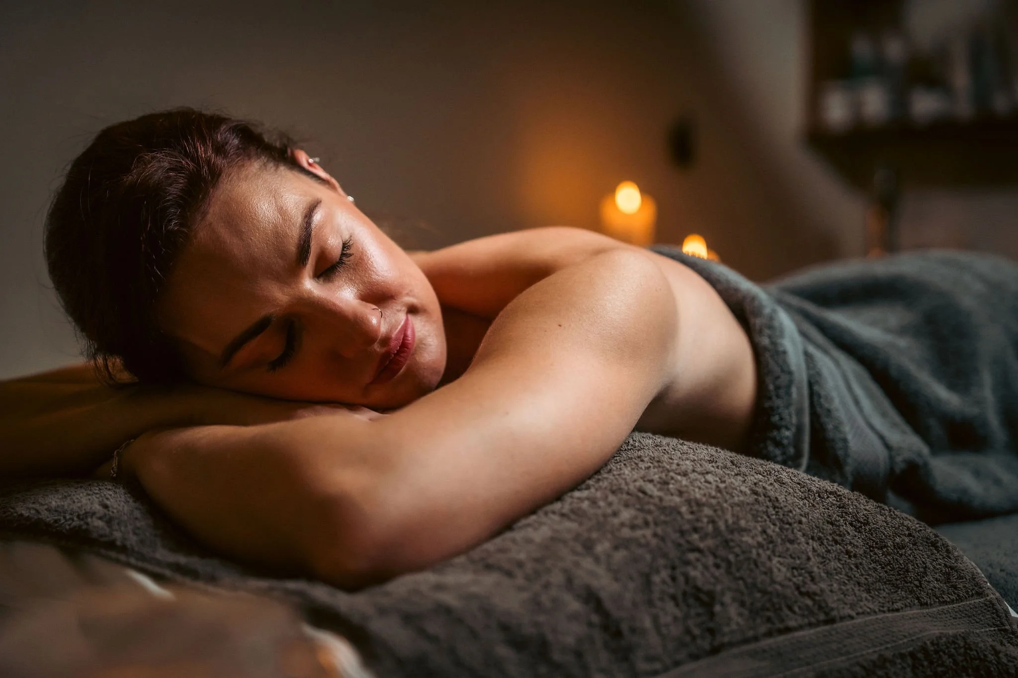 A woman with brown hair relaxing on a massage table in a spa, surrounded by candles.