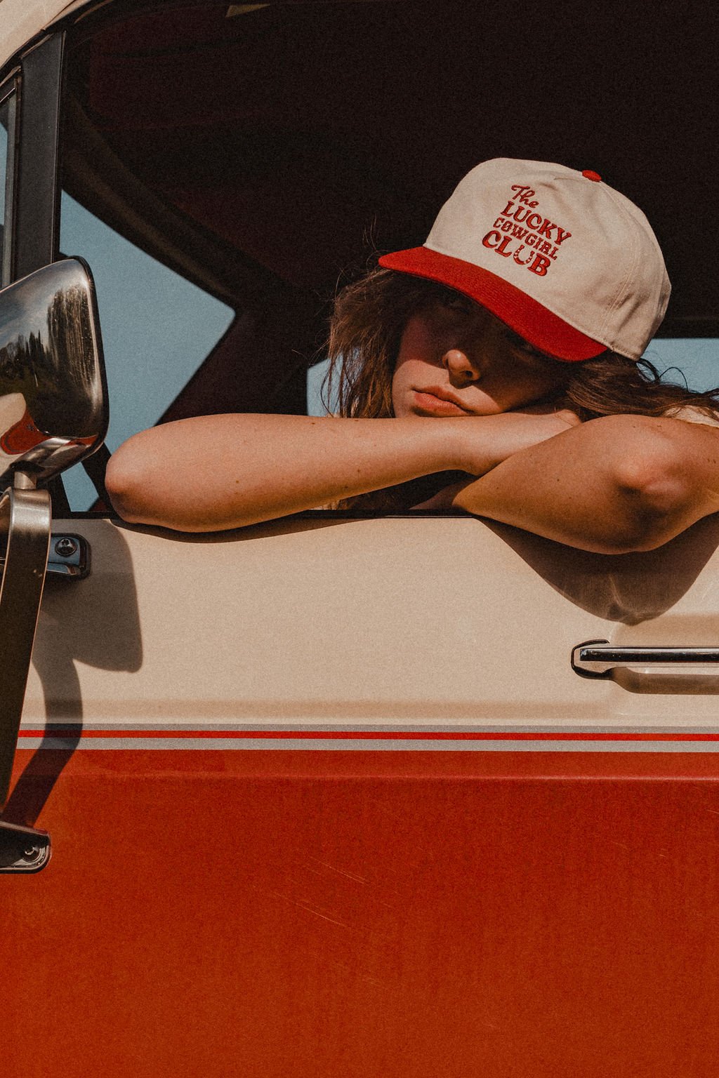 A woman leaning on the window of a vintage van, wearing a baseball cap that reads 'The Lucky Cowgirl Club', with her eyes closed and arms crossed on the window ledge.