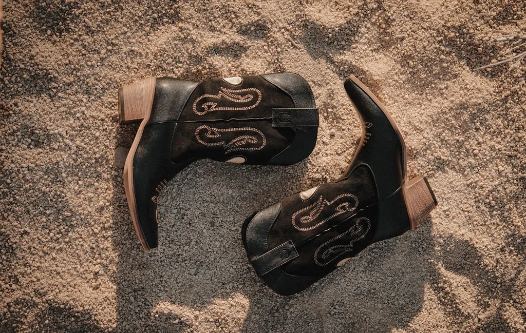 A pair of black cowboy boots with decorative stitching on a sandy surface.