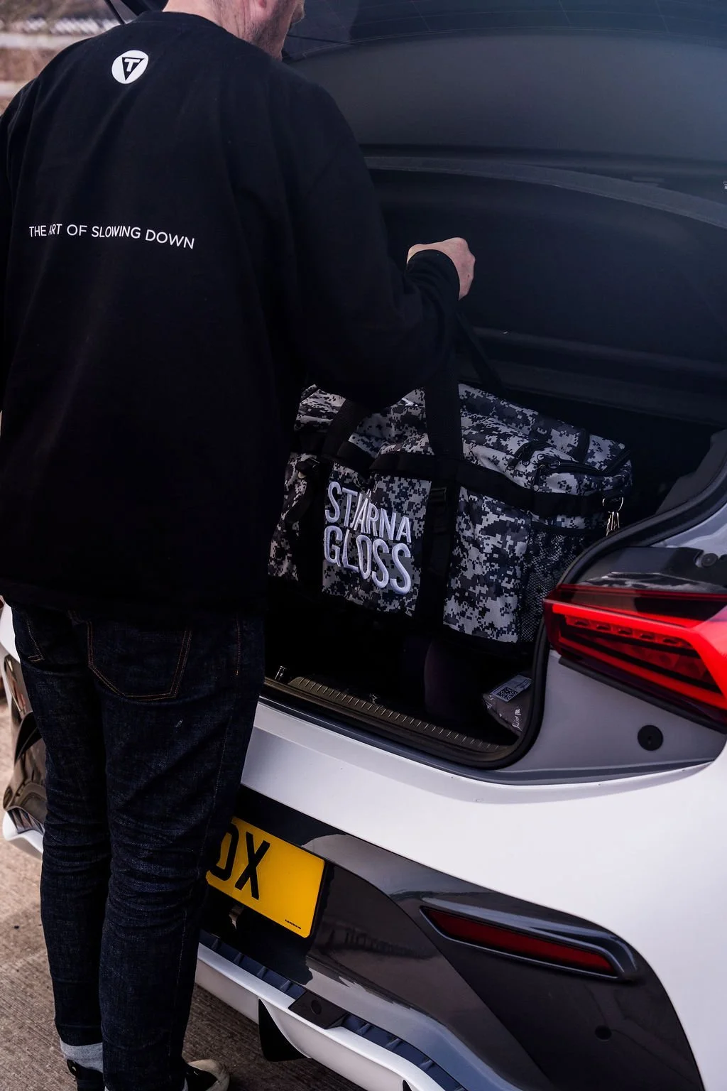 Person loading a camouflage patterned duffel bag with the words "STARNAR GLOSS" onto the trunk of a white car.