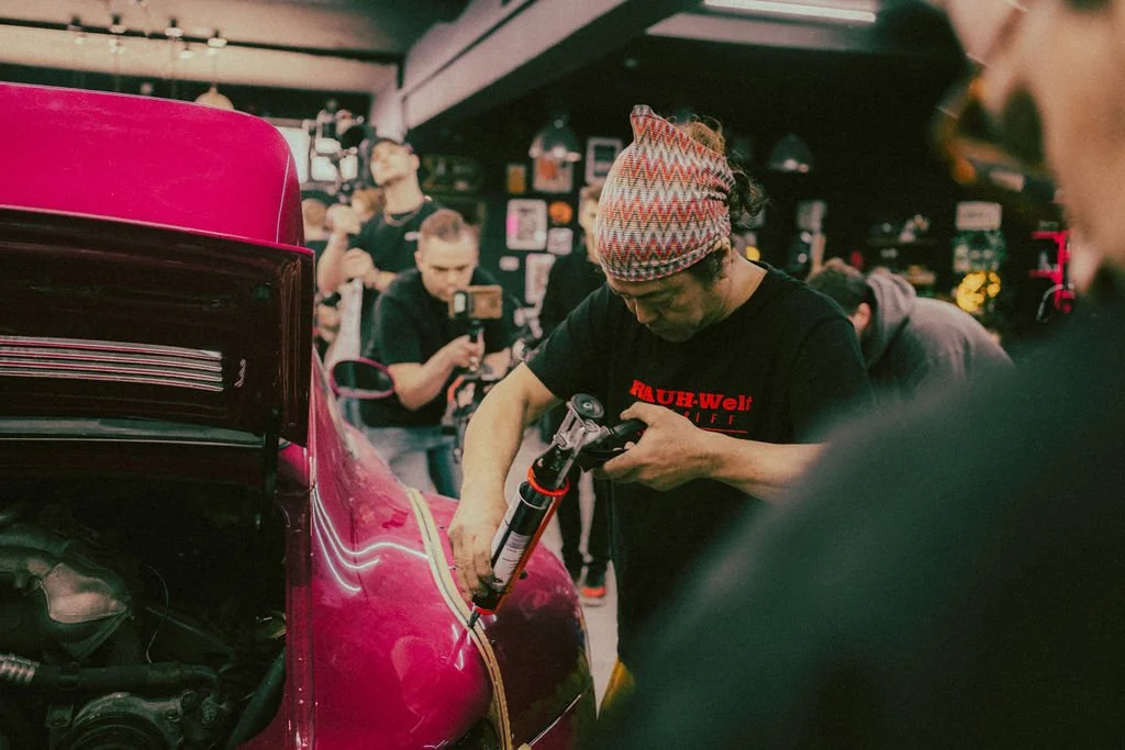 A man in a patterned headband and black T-shirt fixing a red vintage car with a caulking gun inside a garage or workshop, with several onlookers in the background.