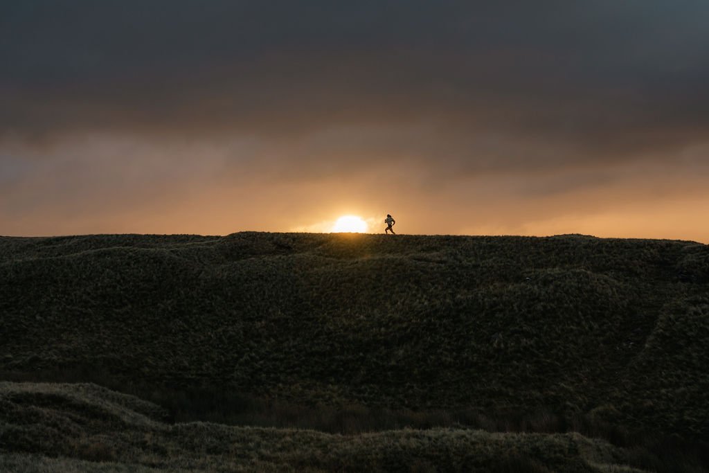 A person hiking along a ridge at sunset with the sun partially set on the horizon, in a natural landscape.