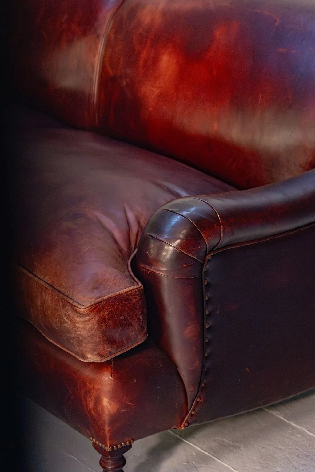 Close-up of a vintage leather armchair with a worn, reddish-brown seat and a dark leather rolled armrest with nailhead trim.