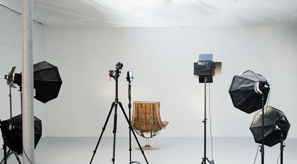 Photography studio with lighting equipment, camera, and a brown chair against a plain white background.
