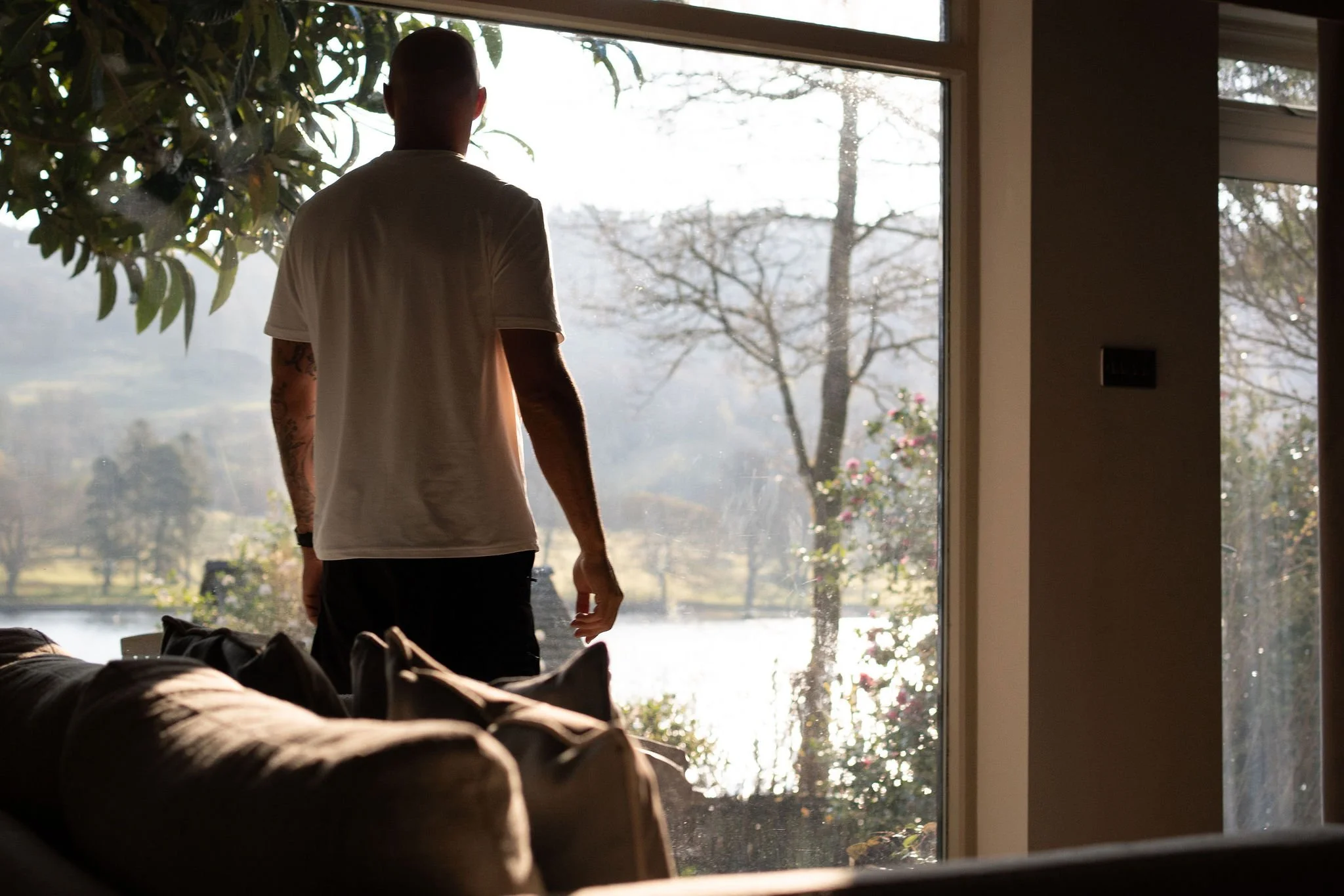 A person with tattoos on one arm, wearing a white T-shirt and dark pants, stands inside a house looking out a large window at a landscape with trees, water, and mountains in the distance during daytime.