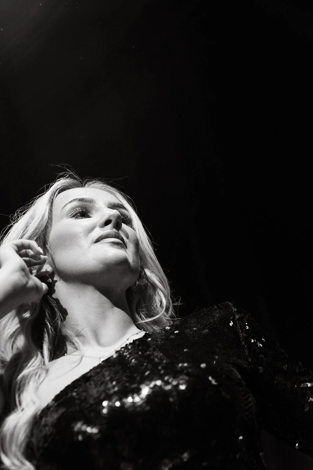 A black-and-white photo of a woman with long, wavy hair, looking upwards with a confident expression. She is wearing earrings and a sequined top, and her hand is near her ear.