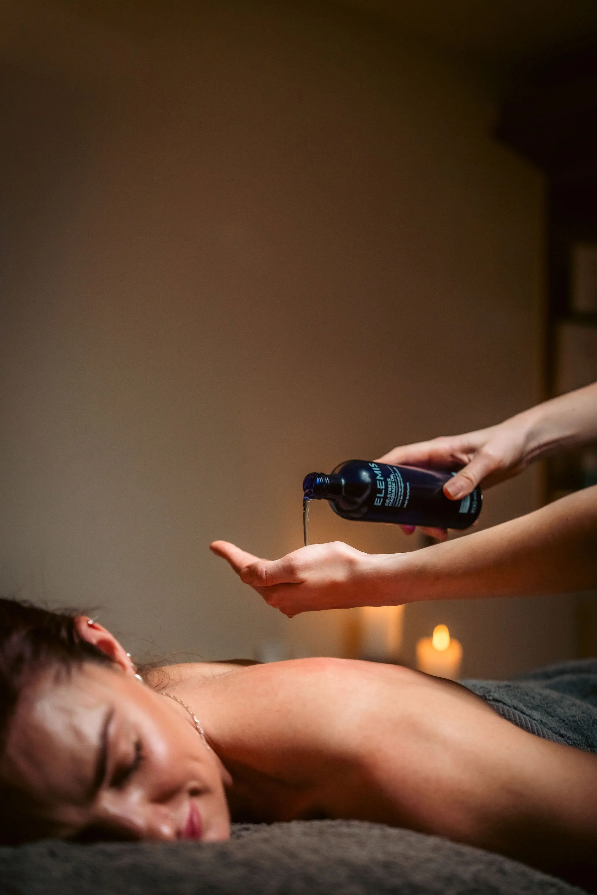 A person receiving a massage with a massage oil being poured onto their back in a relaxing environment with candles in the background.