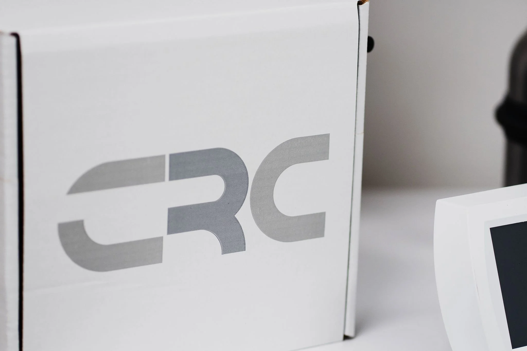 Close-up of a white box with the text 'CRC' on it in grey font, partly obscured, with a small computer monitor or screen to the right.