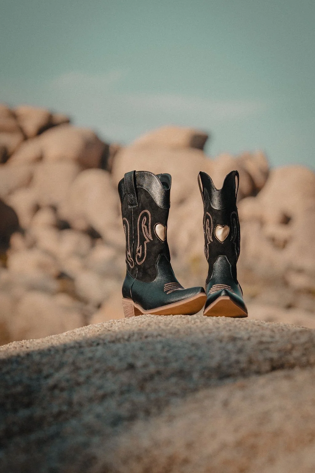 A pair of cowboy boots standing on sandy ground with rocks in the background and a blue sky.