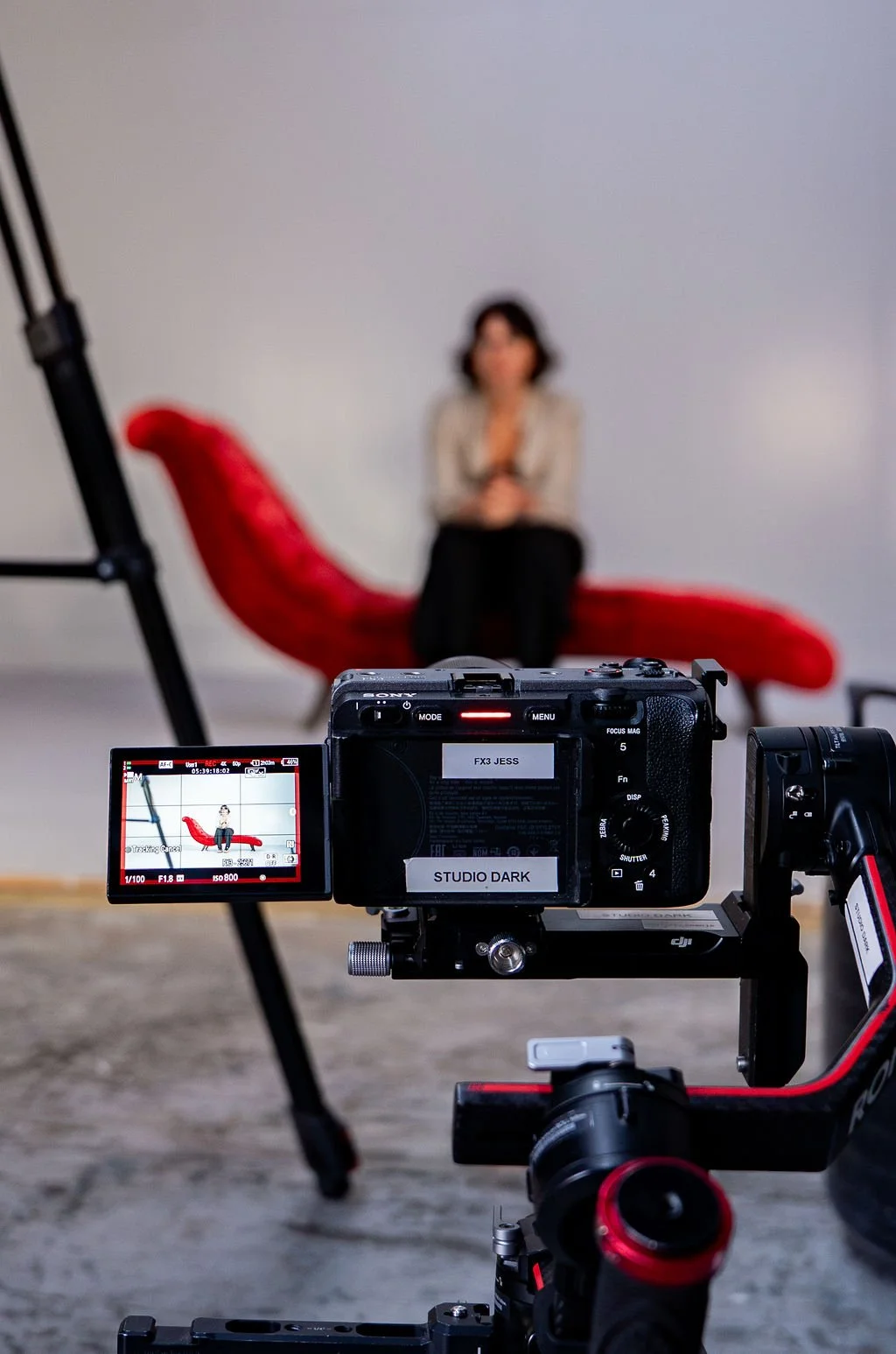 A professional camera on a tripod capturing a woman sitting on a red chaise long, in a photography studio with a plain grey background.